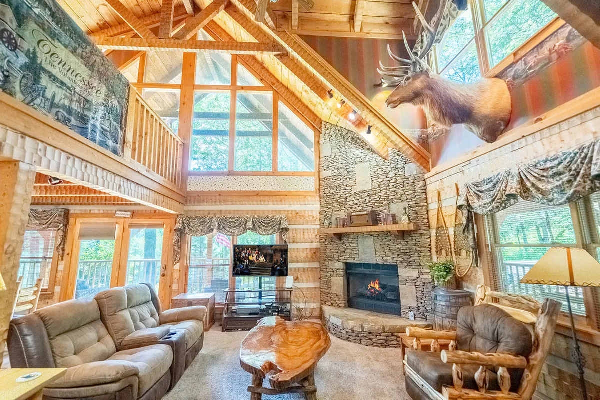 A warm, rustic living room featuring a large stone fireplace, wooden beams, and a unique coffee table surrounded by comfortable seating. Large windows frame views of the surrounding woods, bringing the outdoors in and creating a bright, welcoming space ideal for family gatherings and storytelling.
