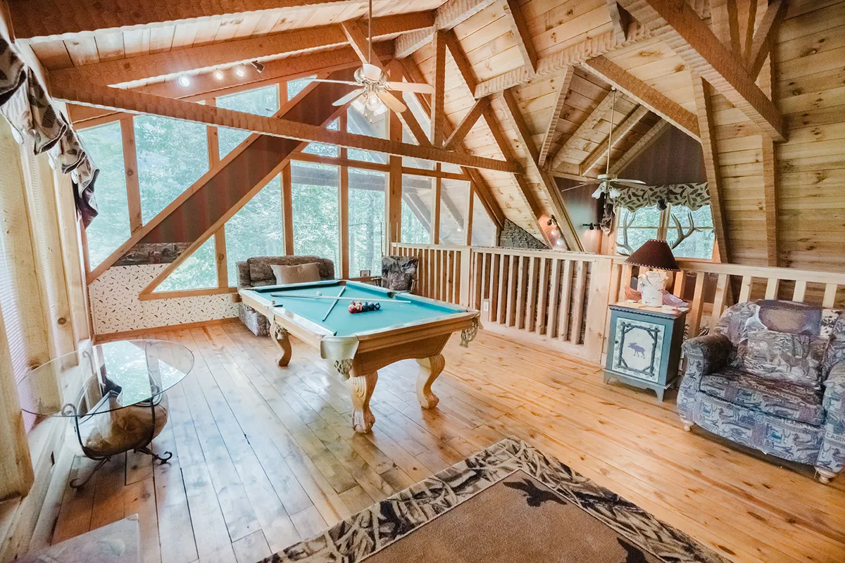 Another view of a loft game room boasting a pool table, comfortable seating, wooden beams, and expansive windows. The relaxed atmosphere is perfect for game nights, reading, or unwinding with friends and family.