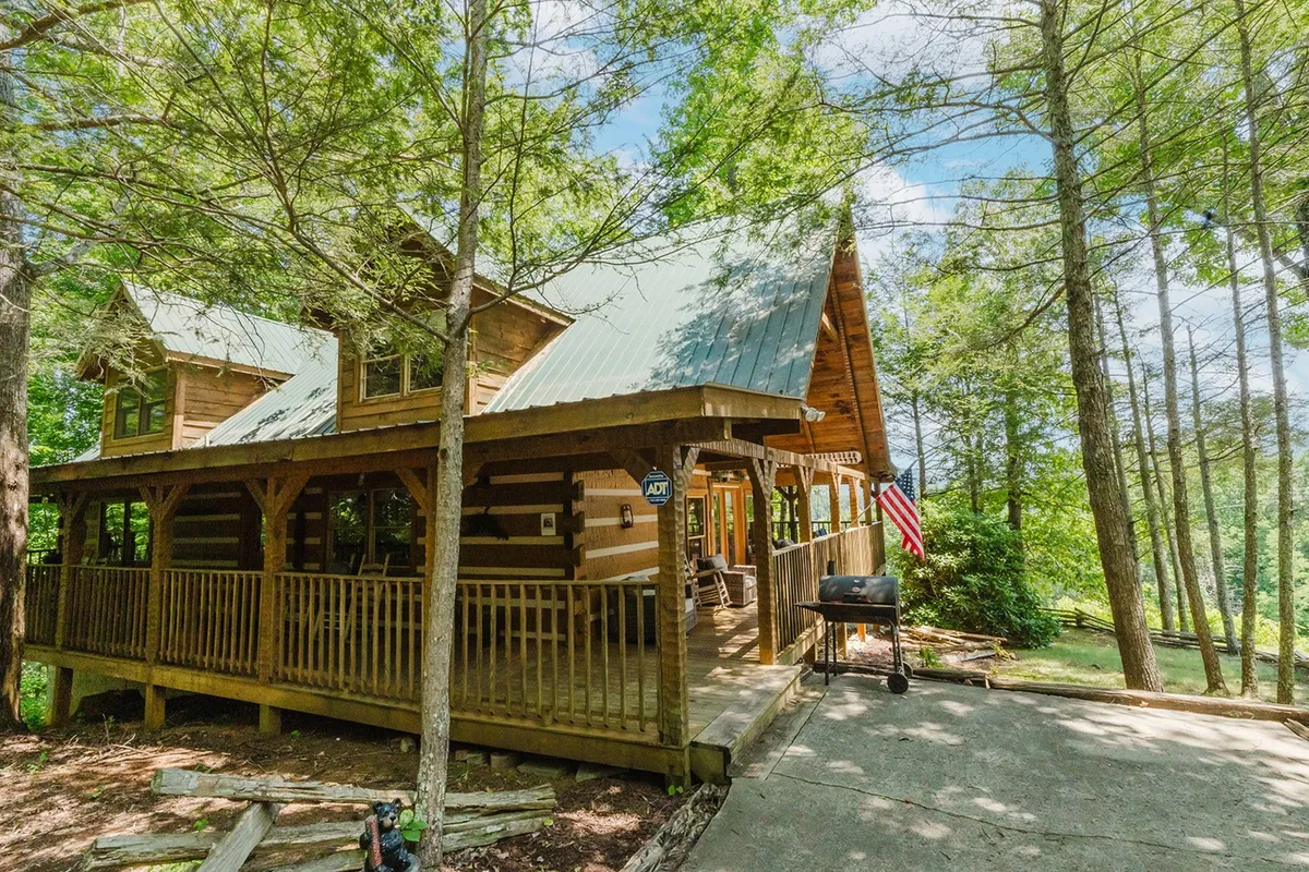 This exterior image showcases the log cabin's charming facade surrounded by lush green trees. The front porch features a grill and an American flag, suggesting a welcoming space for gatherings or relaxing after outdoor adventures. The setting highlights the cabin's natural integration with its environment and hints at easy access to both nature and home comforts.
