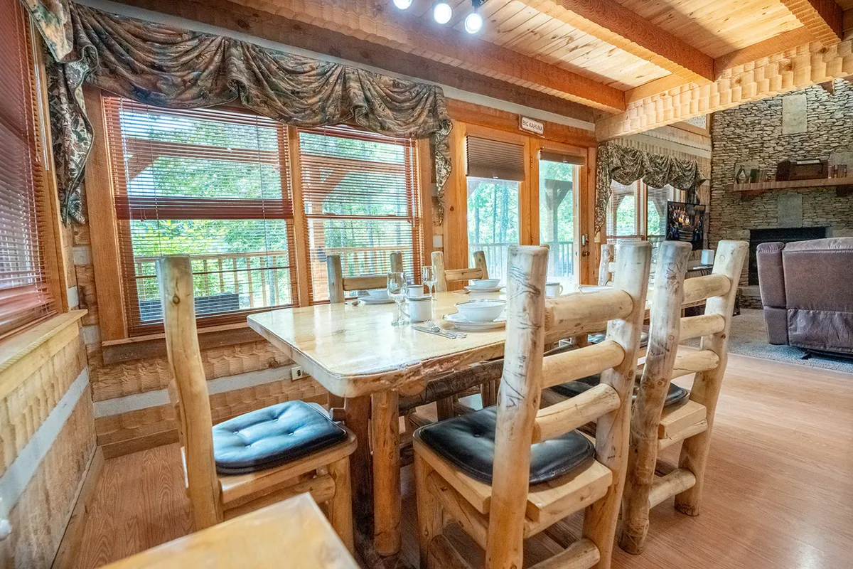 A cozy dining area featuring a wooden table and log-style chairs is set in front of large windows offering a glimpse of the surrounding outdoors. The arrangement invites both casual meals and special gatherings, with space for six and a strong sense of connection to the scenic landscape beyond.