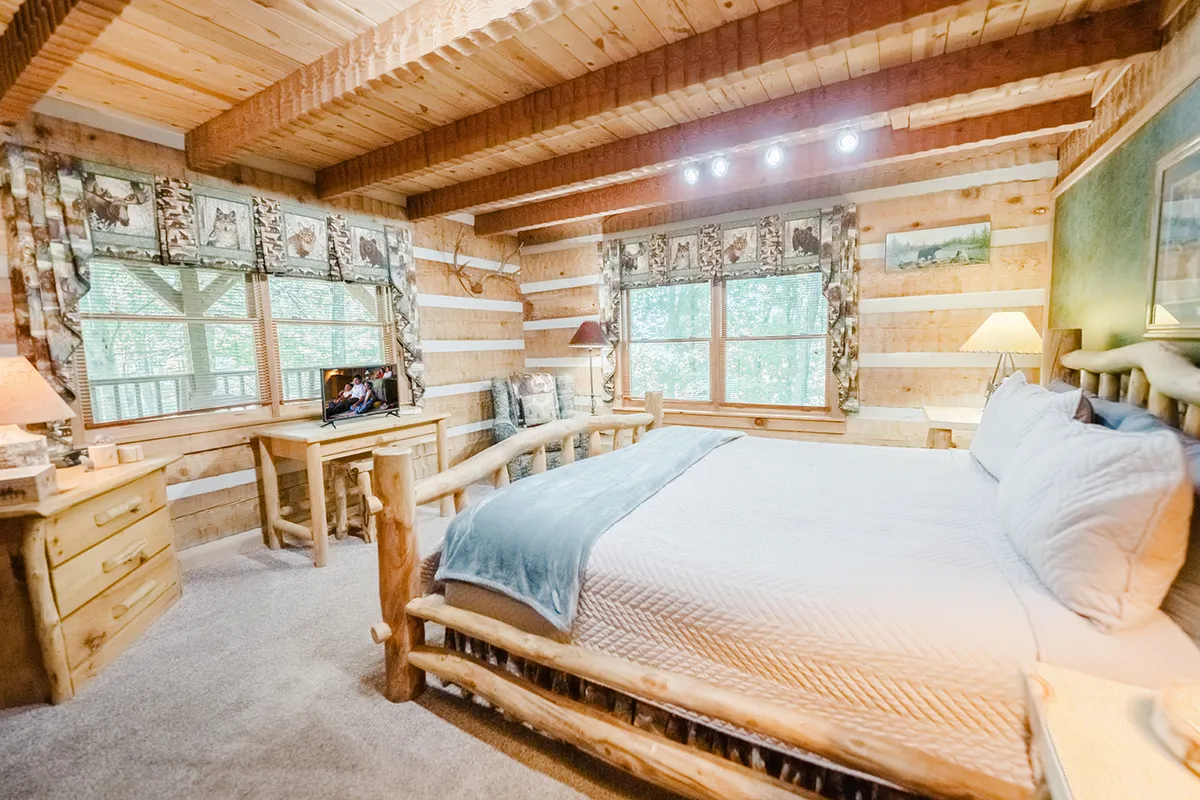 A cozy bedroom with a log bed, wooden furniture, and windows dressed in decorative curtains. The room is bathed in natural light, with spaces for reading by the window, watching TV, or writing at a desk, offering both relaxation and functional touches.
