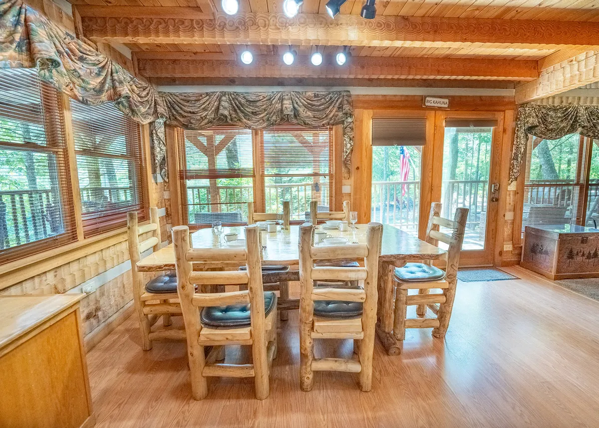 A large wooden dining table surrounded by rustic chairs is showcased in a room filled with natural light from expansive windows. A door leads directly to an outdoor deck, hinting at the cabin's mountain setting. The space feels open, warm, and designed for memorable group meals, games, or celebrations.