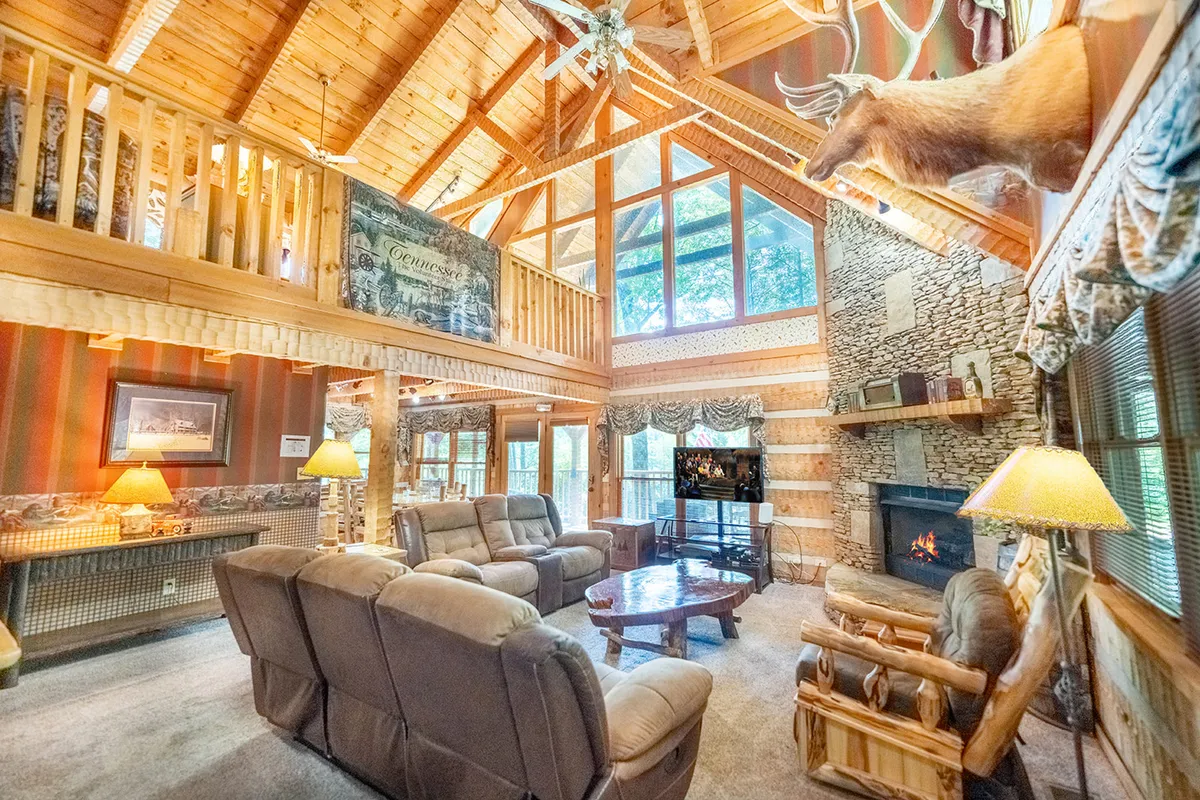 A spacious, open living room with a comfortable seating arrangement centered around a large stone fireplace. Natural light pours in from expansive windows, highlighting the wooden beams and rustic décor. A mounted deer head above the fireplace adds to the cabin's mountain charm, while the inviting sofas and coffee table create an ideal space for relaxation and conversation.