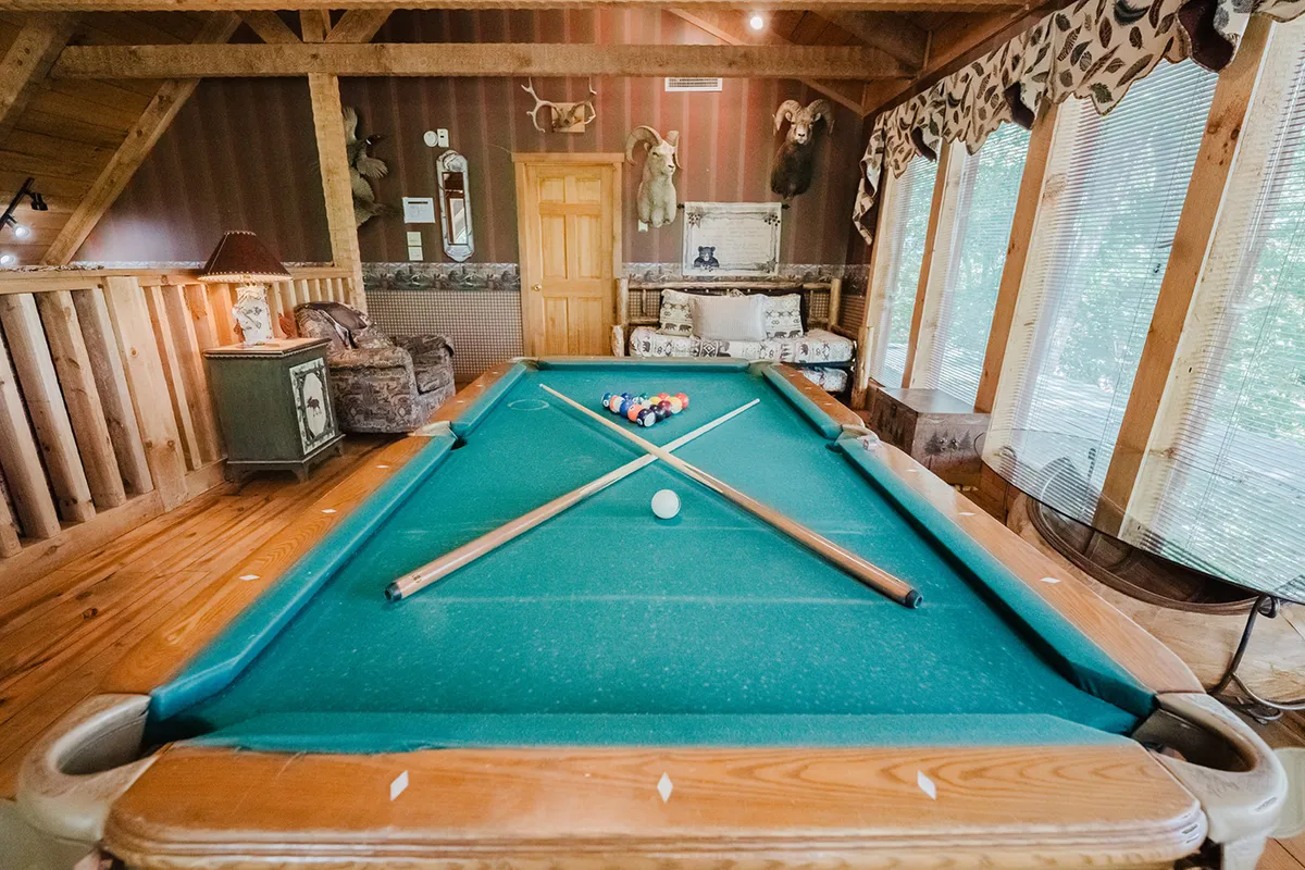 A friendly and relaxed game loft with a pool table at its heart, surrounded by wooden beams, an armchair, and tasteful taxidermy. Large windows brighten the space, making it feel open and inviting. The seating area ensures comfort for spectators or those enjoying quieter activities.