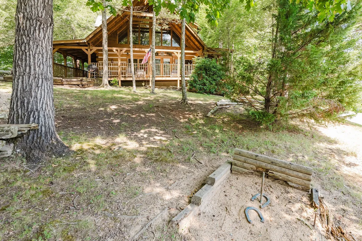 This exterior shot captures a welcoming log cabin with a classic porch, a sand area equipped for horseshoes, and lush greenery surrounding the property. The scene feels vibrant and ready for outdoor activities, suggesting group gatherings and family fun in a natural setting.