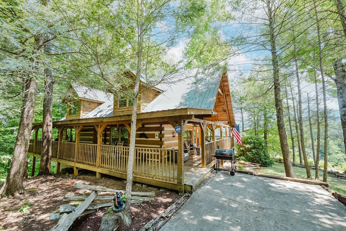 A cozy log cabin exterior featuring a front porch with inviting rocking chairs, a prominent grill, and the American flag amid surrounding trees. The space is ideal for group cookouts, outdoor relaxation, and socializing with friends and family.