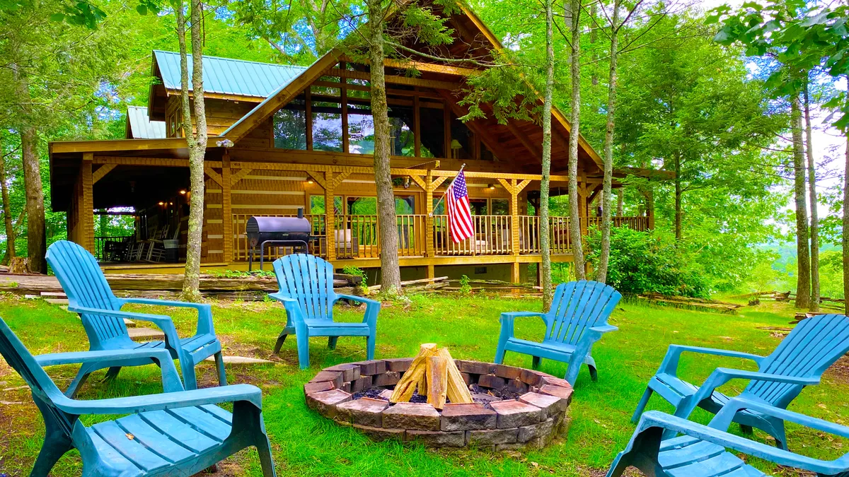 The image showcases the welcoming exterior of a classic log cabin, surrounded by the natural beauty of the Smoky Mountains. In the foreground, a fire pit with blue Adirondack chairs creates an inviting space for guests to gather, with an American flag adding a patriotic, family-friendly touch. The scene exudes relaxation and togetherness, promising memorable evenings spent under the stars.