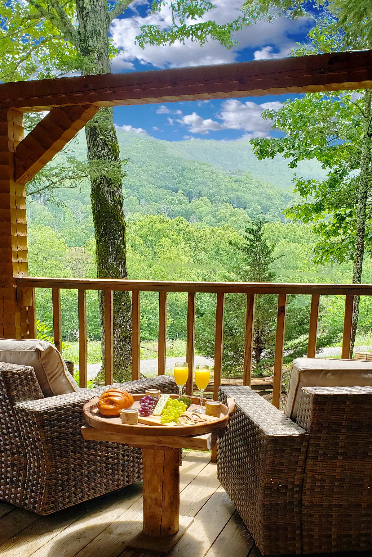 This image showcases a comfortable seating area on a wooden deck with plush chairs and a food platter, all set against a breathtaking backdrop of Smoky Mountain vistas. Sunshine bathes the deck, enhancing the serene environment and inviting guests to relax and savor the view.