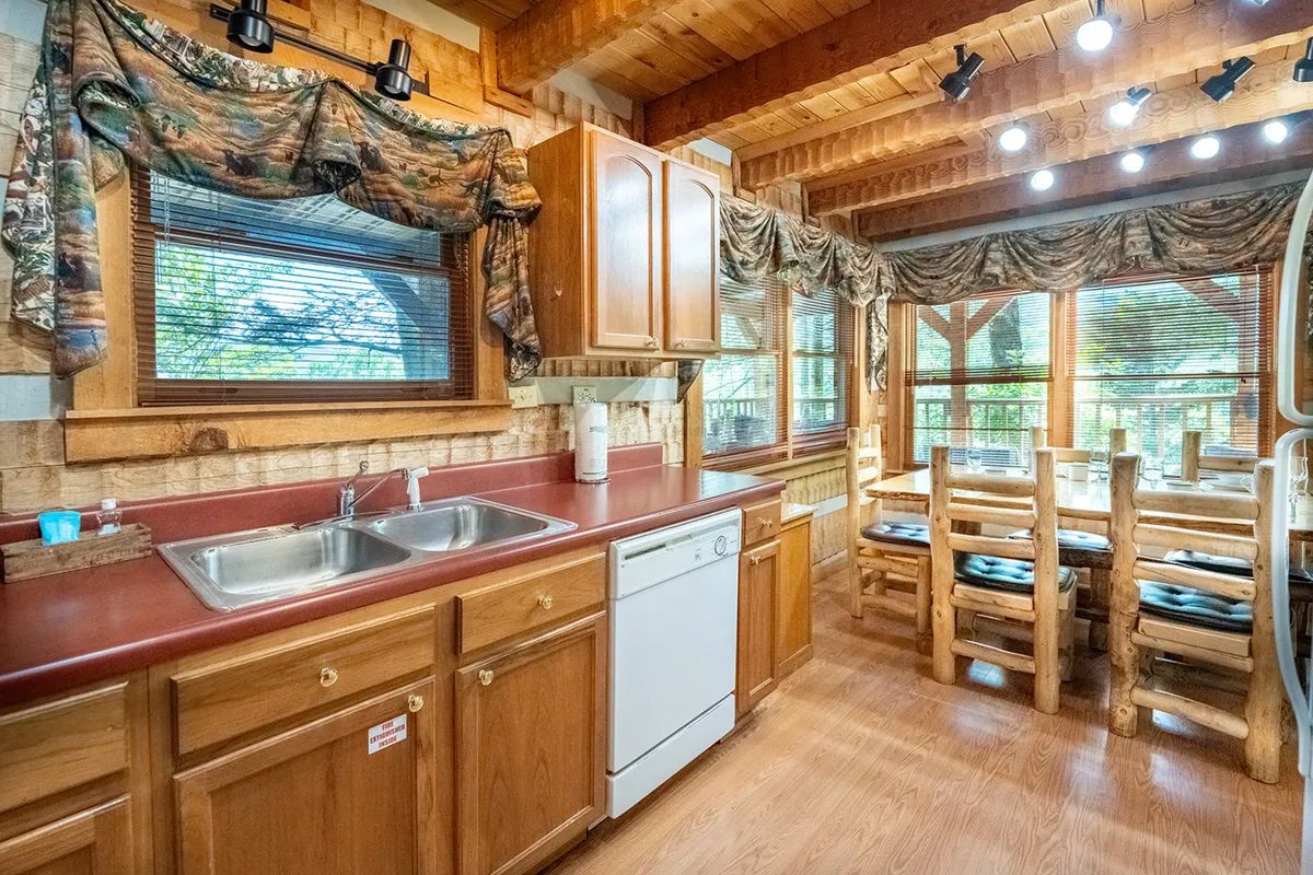 This image presents a welcoming kitchen and dining area with a kitchen counter visible in the foreground and a dining table set near a bright window, capturing a cohesive view of the meal preparation and gathering spaces. Natural light streams in, enhancing the inviting atmosphere. Kitchen appliances and cabinets are neatly arranged, and the dining table is ready for a family meal.