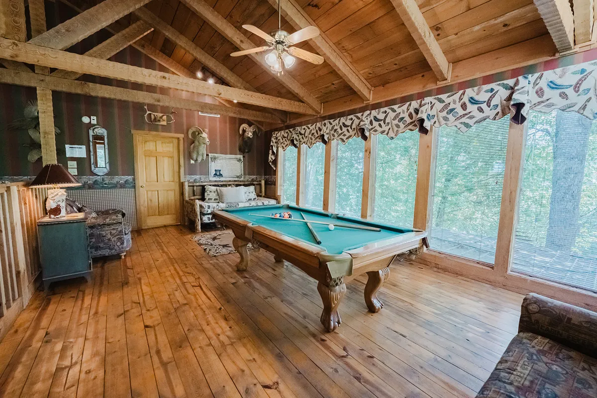 A bright loft game room featuring a pool table as the centerpiece, framed by large windows that let in plenty of natural light. Wooden ceiling beams add rustic charm, while a comfortable sofa seating area encourages lounging and socializing. The overall vibe is cheerful and welcoming, ideal for games and gatherings.