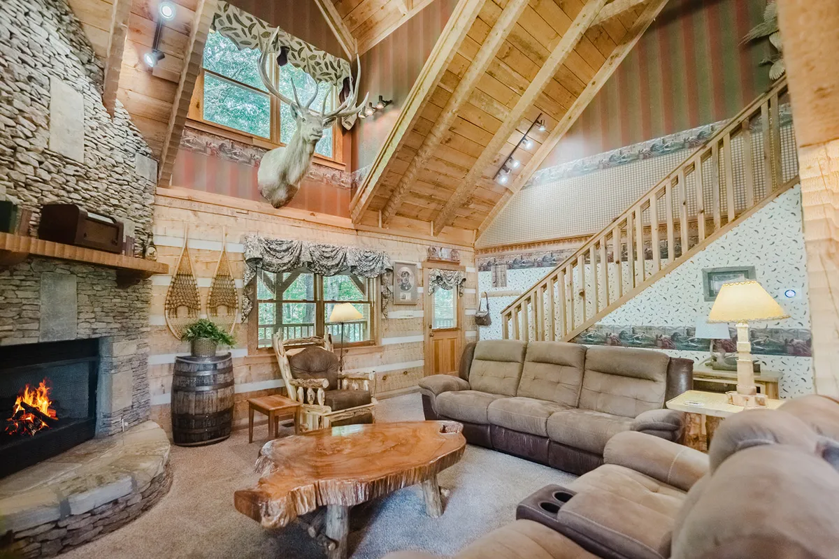 A comfortable, warmly lit seating area with a prominent stone fireplace, large wooden coffee table, and mounted deer head. The layout encourages socializing, with enough space for families and friends to relax together or enjoy a morning coffee with views out the windows.