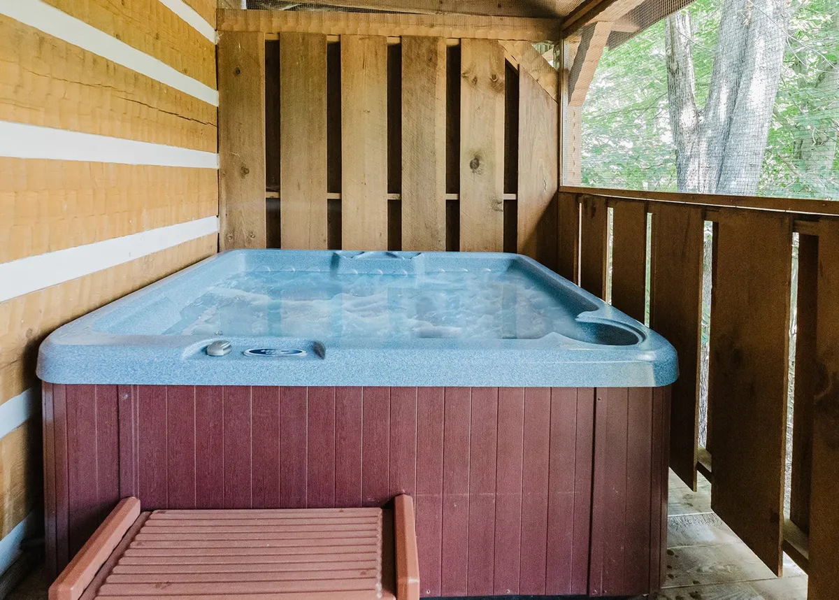 An outdoor hot tub is nestled under a wooden structure surrounded by trees, creating a tranquil ambiance. The scene is ideal for relaxation, with the promise of stargazing or sharing quiet moments while soaking in the warmth of the tub.
